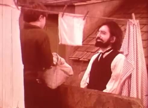 The still from 'Erik Satie and the King of the Beans - Part II' features a young boy interacting with an adult man on a balcony. The adult, presumably inspired by Erik Satie, sports a beard and formal attire. Background elements include hanging laundry and a rustic setting, suggesting a domestic atmosphere. The color palette is muted, creating a nostalgic tone that reflects the film's style.