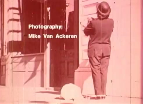 The still features a man, dressed in a suit and bowler hat, standing in front of a building, appearing to engage with the doorway. The image has a distinct pink hue, giving it a stylized, vintage look. Text overlay credits Mike Van Ackeren for photography, suggesting a focus on visual aesthetics in the film 'Erik Satie and the King of the Beans - Part II.' The scene conveys a sense of whimsy and playful engagement with the environment.