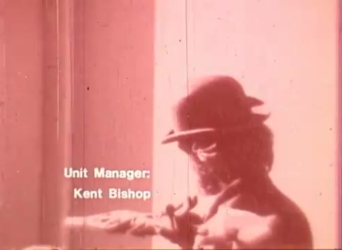 The still from 'Erik Satie and the King of the Beans - Part II' presents a monochromatic image with a reddish tint. It shows a figure wearing a hat, possibly reaching for something or engaged in an action. The credits mention Kent Bishop as the Unit Manager, positioned at the bottom of the frame. The overall aesthetic reflects a vintage film quality, typical of 16mm cinematography.