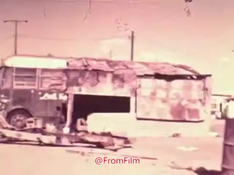 The still from the 16mm print of 'Project Hope' features a dilapidated bus that appears to be partially transformed into a makeshift structure. The surroundings suggest a neglected urban environment, with visible utility poles and a barren landscape. The color tones are muted, typical of vintage film, lending a nostalgic yet somber atmosphere. This scene likely aims to highlight themes of social issues or community struggles prevalent during the 1960s.