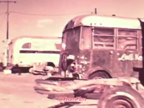 The still from the 'Project Hope' public service announcement features a desolate setting with abandoned vehicles, including a dilapidated bus and trailers in the background. The color palette is muted, typical of 1960s film stock, which conveys a sense of neglect. The environment suggests an urgent social message, likely related to issues faced by communities at the time. Overall, the image encapsulates a somber and reflective tone, aligning with the themes of hope and resilience.