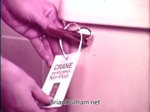 The still features a hand holding a tag attached to a product labeled 'Sani Flush,' which claims to wipe out household germs in 15 seconds. The setting appears to be a bathroom, with a focus on the product and its packaging, highlighting its cleaning capabilities. The color tone is vintage, reflecting the era of the advertisement.