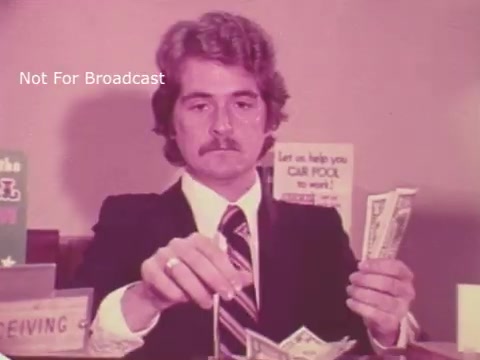 The still from 'Taft for Senate - 1976' features a man with a mustache, dressed in a suit, sitting at a desk. He appears to be handling cash, possibly in a campaign or business setting. The background includes signs that suggest assistance with carpooling, hinting at a community-oriented theme. The overall tone is serious, reflecting a campaign context amidst the political landscape of the time.