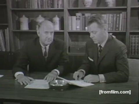 The still from the 1963 'Mystery TV Commercial' features two men seated at a table. They appear to be in a formal setting, possibly an office or studio, with shelves of books and decorative items in the background. One man has a serious expression, while the other appears to be focused on a document in front of him. Both are dressed in suits typical of the era, suggesting a professional or authoritative context, likely related to a medical theme given the mention of the American Medical Association (AMA).  For insights from lip readers, it's best to consult experts in that field, as they can provide accurate interpretations based on visual cues.