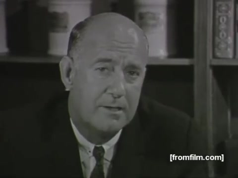 The still from the 'Mystery TV Commercial - 1963' features a middle-aged man in a suit, seated in front of a bookshelf filled with containers. He appears to be speaking directly to the camera, likely delivering a message related to health or medical advice, possibly connected to the American Medical Association. Without sound, the specific content of his speech remains unknown, but his serious demeanor implies that the topic is important. The vintage style of the film reflects the era's communication methods in advertising or public service announcements.