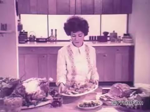 The still features a woman in a retro kitchen, dressed in a white outfit with a decorative pattern. She is carefully holding a platter of food, likely showcasing a meal, while two large roasted dishes are placed nearby. The kitchen has a mid-century modern aesthetic, with wooden cabinetry and a bright atmosphere, typical of the 1960s. This context suggests a promotional scene for the Reveal See-Through Roasting Wrap, highlighting convenience and presentation in cooking.