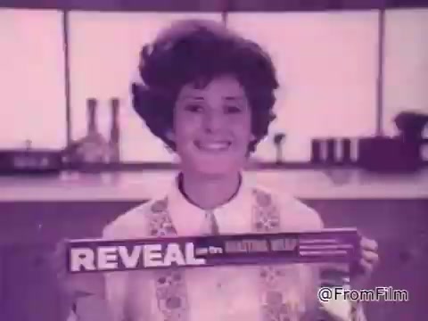 The still features a woman with a stylish 1960s hairstyle, smiling as she holds a box of 'Reveal See-Through Roasting Wrap.' The background suggests a kitchen setting, with soft lighting and kitchen utensils visible, enhancing the retro feel of the advertisement. The overall tone is cheerful and promotional, highlighting the product's appeal.