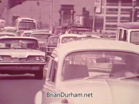 The still from the 'Iron City Beer' commercial shows a busy street scene filled with vintage cars in heavy traffic. The overall tone is nostalgic, capturing a moment likely from the mid-20th century. The cars, predominantly in pastel colors, reflect the era's design, while the urban backdrop suggests an industrial setting, typical of Pittsburgh. The ambiance hints at a lively city life, possibly promoting Iron City Beer as part of the local culture.