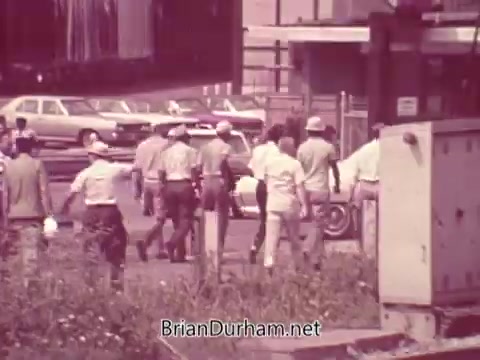 The still from the 'Iron City Beer' commercial depicts a group of individuals in casual attire, potentially walking through an industrial or urban area. In the background, parked cars suggest a mid-20th century setting, and nearby foliage indicates some green space amidst the concrete. The scene conveys a lively, communal atmosphere, typical of beer advertisements from that era, focusing on camaraderie and social enjoyment.
