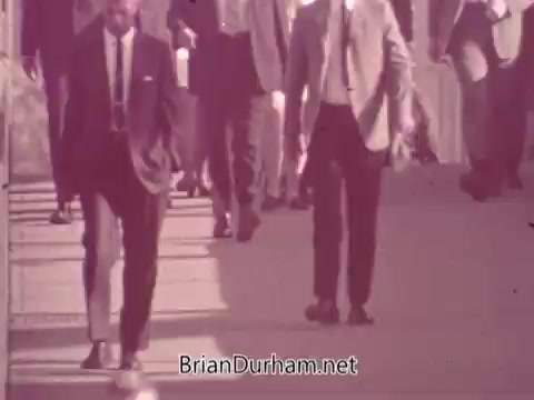 The still features a group of well-dressed men in suits walking along a city sidewalk. The scene captures a bustling urban environment typical of mid-20th century America, portraying a sense of camaraderie and social interaction. The mood is casual yet sophisticated, reflecting the culture surrounding Iron City Beer during that era.