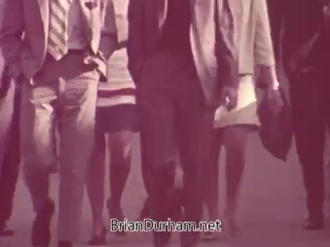 The still from the 'Iron City Beer' commercial features a group of people walking in a city setting. They are dressed in formal attire, indicating a professional or polished atmosphere. The image conveys a sense of camaraderie and urban life, typical for beer marketing, emphasizing social connections. The color tone suggests a vintage style, characteristic of older advertising campaigns, enhancing the nostalgic feel.