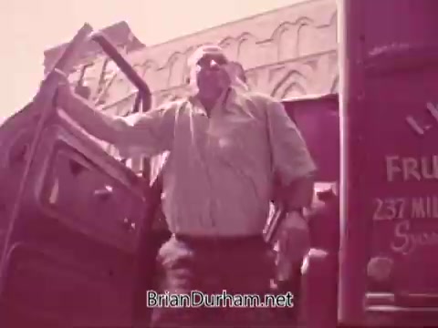 The still from the 'Iron City Beer' commercial features a muscular man exiting a delivery truck, likely depicting a scene of local beer distribution. The colors are warm, with a vintage aesthetic typical of 16mm film. The setting indicates a bustling urban environment, suggesting a connection to the community and the promotional spirit of the brand.