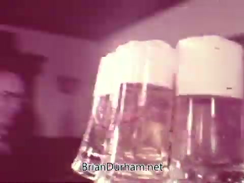 The still from the 'Iron City Beer' commercial features three glasses of beer, each with a frothy white head. The background suggests a social setting, likely a bar or gathering place. The focus is on the glasses, emphasizing camaraderie and celebration associated with the brand. The image has a vintage feel typical of 16mm film.