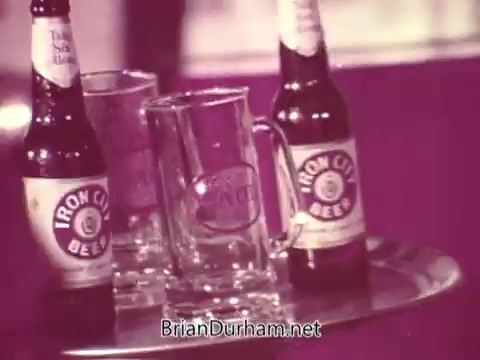 The still features two bottles of Iron City Beer alongside two empty pint glasses on a silver tray. The branding is clearly visible on the bottles and glasses. The composition suggests a casual drinking setting, likely indicating a social atmosphere in a promotional context. The overall tone is warm and inviting, representative of classic beer advertising.