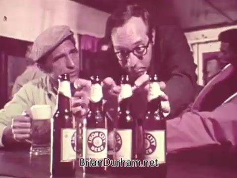 The still features two men seated at a bar, focusing on several bottles of Iron City Beer placed in front of them. One man appears to be gesturing enthusiastically about the beer, while the other listens attentively. The scene captures a casual, social atmosphere typical of beer commercials. The branding on the bottles is prominently displayed, emphasizing the product.