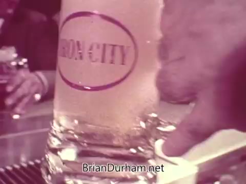 The still from the Iron City Beer commercial features a close-up of a frosty glass of beer with the 'Iron City' logo prominently displayed. A hand is shown reaching towards the glass, suggesting service or a toast. The setting appears to be a bar with other patrons in the background, creating a social atmosphere. The image has a vintage look, characteristic of mid-20th century advertisements.