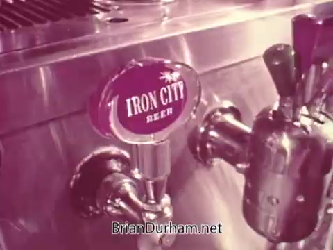 The still features a bar tap with an 'Iron City Beer' handle prominently displayed. The tap is mounted on a shiny metal surface, showcasing the bar's polished chrome fixtures. The aesthetic suggests a vintage or retro style, consistent with a mid-20th century commercial vibe.