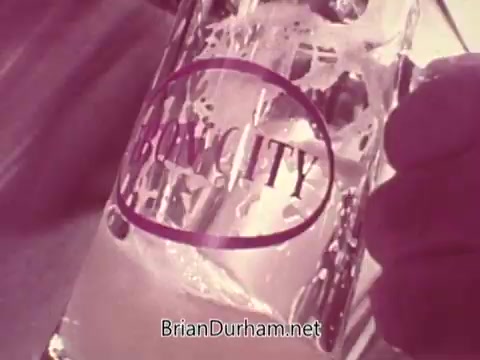 The still features a close-up of a glass with the branding 'IRON CITY' prominently displayed. The beer appears light in color, with visible bubbles and condensation on the glass, suggesting it's cold and refreshing. The overall tone suggests a nostalgic or retro aesthetic associated with classic beer commercials.