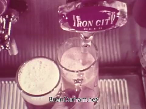 The still features a close-up of a bartender pouring Iron City Beer from a tap into a glass. The beer has a frothy head, and the logo for Iron City is prominently displayed on the tap handle. The setting suggests a bar environment, emphasizing the pouring process and the beer's quality.