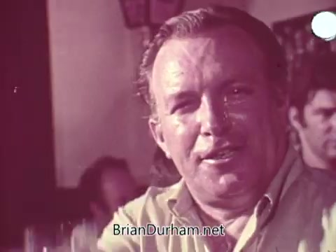 The still from the 'Iron City Beer' commercial features a man sitting at a bar, smiling while engaging with the camera. The atmosphere suggests a casual and friendly environment, with other patrons visible in the background. The focus is on the man's expression, promoting a sense of enjoyment associated with the beer. The overall aesthetic is reminiscent of vintage advertising from the mid-20th century.