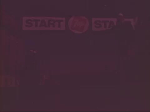 The still from the 1978 7-Up commercial features a dimly lit scene with the word 'START' prominently displayed on a banner. The image suggests a dynamic setting, possibly related to a competition or event, but lacks clarity due to low visibility. The overall atmosphere evokes excitement and anticipation typical of a promotional ad.