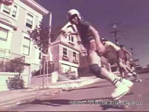 The still shows a young skater wearing a helmet and protective gear, gliding down a sloped street lined with residential houses. The visual style reflects the 1970s, featuring vibrant colors and a sense of youthful adventure. Other skaters are in the background, contributing to a fun, energetic atmosphere typical of the era's advertisements. The overall scene captures a sense of freedom and the joy of outdoor activities, aligning with the promotional spirit of the 7-Up brand at the time.