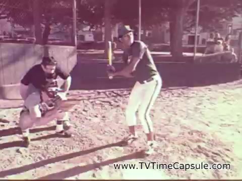 The still depicts a scene from a 1978 7-Up commercial featuring two young boys on a baseball diamond. One boy is in a catching stance, wearing a cap, while the other is poised to bat. The setting is casual, with a dirt field and trees in the background, capturing a nostalgic vibe typical of the era.