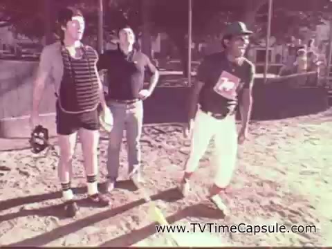 The still features three men in a casual outdoor setting, likely a baseball field. Two of the men are dressed in sporty attire, with one wearing a striped shirt and shorts, and the other in a T-shirt and white pants. The third man is in a cap and appears to be preparing to throw a baseball. The scene conveys a relaxed, playful atmosphere typical of a 1978 7-Up commercial, emphasizing enjoyment and camaraderie.