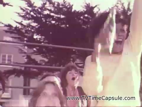 The 16mm still from the 1978 7-up commercial captures a lively outdoor scene with people joyfully celebrating. Individuals are seen cheering and expressing excitement, with one person in a yellow shirt raising their arms triumphantly. The backdrop features trees and a casual gathering atmosphere, emphasizing a sense of fun and celebration associated with the beverage.