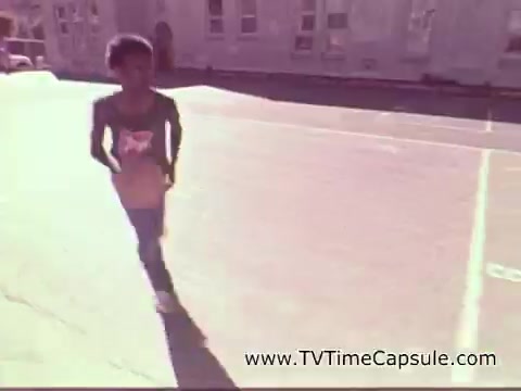 The still from the 1978 7-Up commercial features a young boy running on a sunlit street, showcasing a playful and energetic vibe. He wears a T-shirt that likely promotes identity or fun, capturing the carefree spirit of childhood in that era. The background shows a residential setting, contributing to a nostalgic atmosphere.