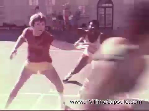 The still from the 1978 7-Up commercial features two male athletes engaged in an outdoor game, likely basketball. One player, wearing a red shirt and yellow shorts, is in a dynamic stance, seemingly on defense. The scene captures a sense of action and energy, characteristic of sports advertisements of the era, promoting an active lifestyle alongside the refreshing drink. The background includes outdoor settings, adding to the lively atmosphere.
