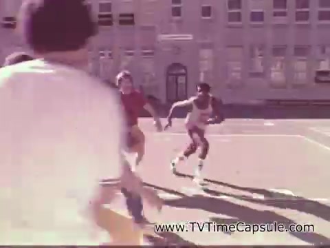 The still shows a group of young people playing basketball outdoors, likely on a school playground or court. One player in a tank top is in motion, while others are engaged in the game, creating a dynamic and energetic atmosphere. The scene evokes a sense of youth and fun, aligning with the nostalgic vibe of a 1978 7-Up commercial. The setting appears sunny, suggesting an enjoyable day for outdoor activities.