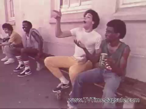 The still from the 1978 7-Up commercial shows a group of young men sitting on a bench. One individual in a white shirt is animatedly talking or laughing, while another next to him holds a can of 7-Up. The setting has a retro vibe, typical of the late 1970s, featuring casual clothing and a relaxed atmosphere. The overall tone suggests camaraderie and enjoyment.