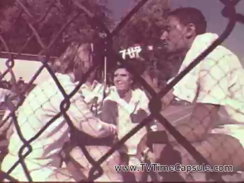 The still features three people interacting passionately, likely in a playful or competitive setting. The foreground shows two individuals, one male and one female, while another male is positioned off to the side. They wear white clothing, and a sign for 7-Up is visible in the background, suggesting that they are promoting the beverage. The scene is framed by a wire fence, adding a sense of context to the social or sporting atmosphere of the commercial. The overall tone appears lively and engaging, characteristic of 1970s advertising.