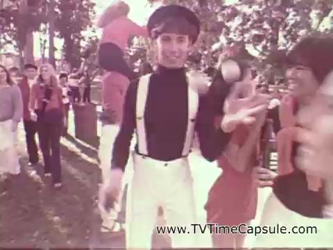 The still from the 1978 7-Up commercial features a lively, outdoor scene with a group of young people. A central figure, dressed in a black turtleneck and white pants with suspenders, smiles while juggling. Surrounding him, others, dressed in colorful outfits, appear to be enjoying themselves, contributing to a festive and youthful atmosphere. The overall vibe is upbeat and playful, reflecting the brand's fun image.