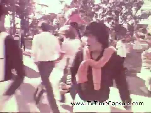 The still from the 1978 7-Up commercial features a lively outdoor scene. A person in focus is wearing a hat and a bright orange scarf, looking animated as they interact with the crowd around them. People in the background are walking, creating a bustling atmosphere typical of a fun, leisurely day. The overall vibe captures a sense of enjoyment and refreshment associated with the beverage.