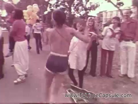 The still from the 1978 7-Up commercial features a lively outdoor scene. A young person in shorts is dancing enthusiastically, surrounded by a diverse group of people enjoying a gathering. Balloons and a carefree atmosphere suggest a festive event, with onlookers smiling and engaging with the performer. The setting captures a sense of fun and spontaneity typical of commercial advertisements from that era.