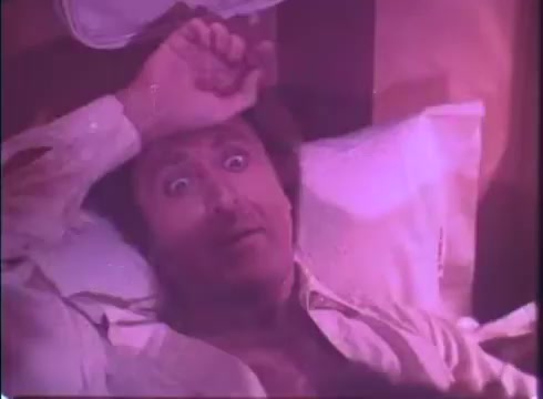 The still from the movie trailer 'Silver Streak' captures a character in a bed, looking alarmed with wide eyes and a hand raised to his forehead, suggesting a moment of surprise or realization. The scene is lit with a purple hue, contributing to an intense or comedic atmosphere typical of the film’s blend of comedy and suspense. The character is dressed in a slightly disheveled manner, indicating a state of chaos or confusion.