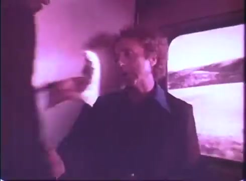 The still from the 'Silver Streak' trailer features a tense scene set inside a train. A man with curly hair is seated, looking alarmed as a shadowy figure approaches him with an outstretched hand, possibly threatening. The window behind them shows a blurred landscape, indicating motion, enhancing the suspenseful atmosphere of the moment. The overall color tone is slightly purple, indicative of the 16mm film quality, adding a vintage feel to the scene.