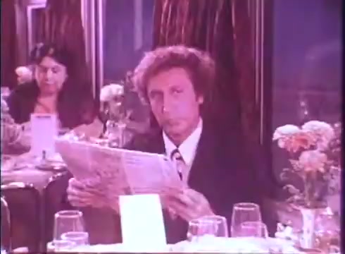 The still depicts a scene from the movie trailer for 'Silver Streak,' showcasing a man in a suit seated at a table in a restaurant. He is reading a newspaper, exhibiting a contemplative expression. The background features a woman and floral arrangements, contributing to an elegant dining atmosphere. The overall tone suggests a moment of intrigue or tension within the narrative.