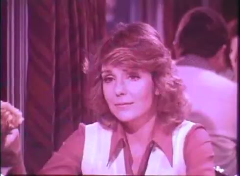 The still features a woman seated at a table in a restaurant, with a soft focus and a slight pink hue typical of 16mm prints. She has wavy, shoulder-length hair and a contemplative expression, wearing a collared blouse. The background suggests a busy diner atmosphere, hinting at conversation and activity, possibly capturing a moment of intrigue or connection in the trailer for 'Silver Streak.'