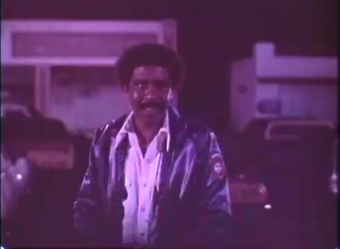 The still from the movie trailer 'Silver Streak' features a man wearing a blue jacket with a white shirt underneath, standing outdoors at night. He has a smile on his face and is likely in a parking lot setting, with blurred cars and a faint backdrop indicating a nighttime city atmosphere. The overall tone appears light-hearted and engaging, typical of a comedic adventure film. The image quality reflects the vintage nature of a 16mm print, giving it a nostalgic feel.
