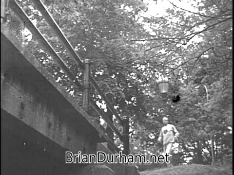 The still shows Ray Bolger, known for his role as the Scarecrow in 'The Wizard of Oz,' in a public service announcement for the Heart Fund from the 1960s. He is depicted walking along a path under a bridge surrounded by trees, conveying a message about heart health. The black-and-white format adds a nostalgic feel typical of the era.