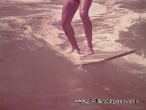The 16mm still from 'Army Recruiting Commercial - Your Man in Waikiki' depicts a surfer gliding on a surfboard through the ocean waves. The focus is on the surfer's legs and the surfboard, highlighting the dynamic motion and the vibrant beach environment of Waikiki. The scene conveys a sense of freedom and adventure, aligning with the promotional theme of the commercial.