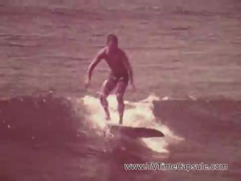 The still from the 'Army Recruiting Commercial - Your Man in Waikiki' shows a surfer catching a wave. The scene captures a sunny, vibrant beach setting typical of Waikiki, emphasizing leisure and an active lifestyle. The surfer, likely a young man, is dressed in swim trunks and appears confident as he balances on the board, showcasing skill and enjoyment in the ocean, linking the image of military service with adventure and an idealized, energetic atmosphere.