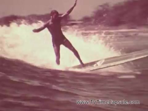 The still from the 'Army Recruiting Commercial - Your Man in Waikiki' shows a surfer in action, riding a wave. The surfer is dressed in a wetsuit and appears dynamic as they maneuver on the surfboard. The backdrop features ocean waves and a coastal landscape, emphasizing a vibrant, outdoor lifestyle. The overall tone suggests a sense of adventure and freedom, aligning with the recruitment theme of showcasing life in Hawaii.