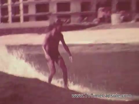 The still depicts a man surfing in Waikiki, capturing a vibrant and energetic atmosphere. He is dressed in a swimsuit, balancing on a surfboard as he rides a wave, showcasing the thrill of the sport. The background features sandy shores and hints of resort buildings, emphasizing the tropical, recreational setting. The overall tone is nostalgic and promotional, typical of an Army recruiting commercial aimed at engaging viewers with a dynamic lifestyle.