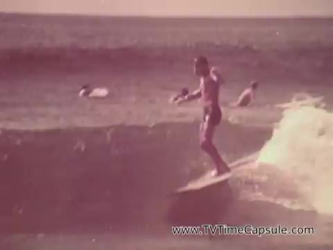 The still features a surfer riding a wave in a sunny, beach setting typical of Waikiki. In the background, other surfers can be seen enjoying the water. The atmosphere is vibrant and leisurely, indicative of a carefree lifestyle associated with the beach scene. This visual aligns with the promotion of Army recruitment, suggesting a connection between service and the appealing aspects of living in such an idyllic environment.