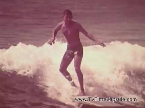 The still from the Army Recruiting Commercial 'Your Man in Waikiki' features a shirtless man surfing on a wave. He appears athletic and confident, embodying a lively beach culture. The hues of the image suggest a vintage aesthetic, typical of 16mm film. The context implies a recruitment message, connecting the fun and adventurous lifestyle of surfing in Waikiki with the spirit of military service.