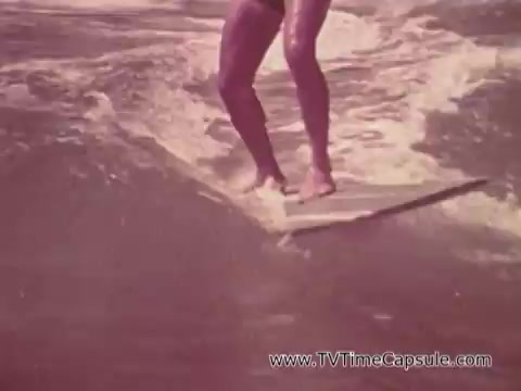The still from the 'Army Recruiting Commercial - Your Man in Waikiki' features a person surfing on a wave. The surfer is barefoot and balances on a surfboard, showcasing a relaxed and carefree lifestyle associated with beach culture in Waikiki. The color tones suggest a vintage aesthetic, typical of 16mm film, enhancing the nostalgic feel of the scene.